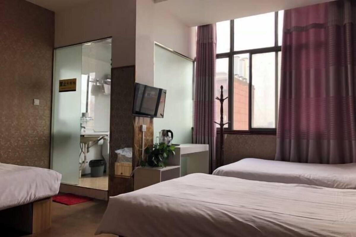 Quarto de hotel com duas camas, televisão, janela com cortinas e vista para um edifício.