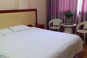 A hotel room with a bed, a bedside table, a chair, a small round table, and a vase with flowers. - Guzhen Zihao Hotel (Guzhen)