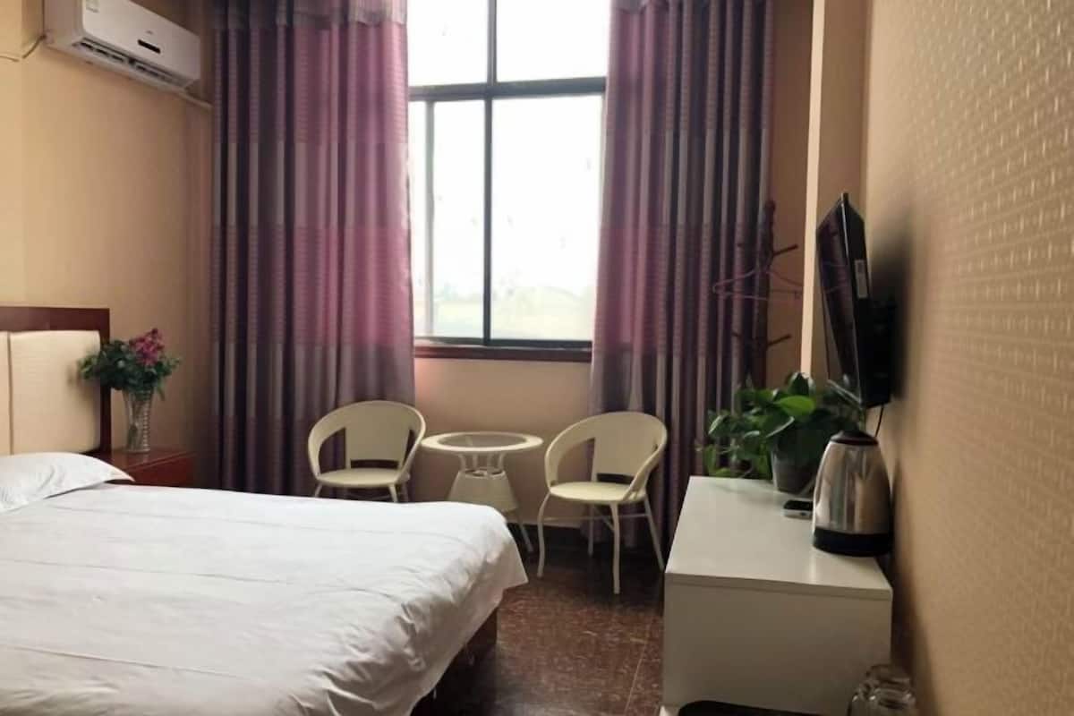 Quarto de hotel com cama, duas cadeiras, uma mesa pequena, televisão e janela com cortinas.