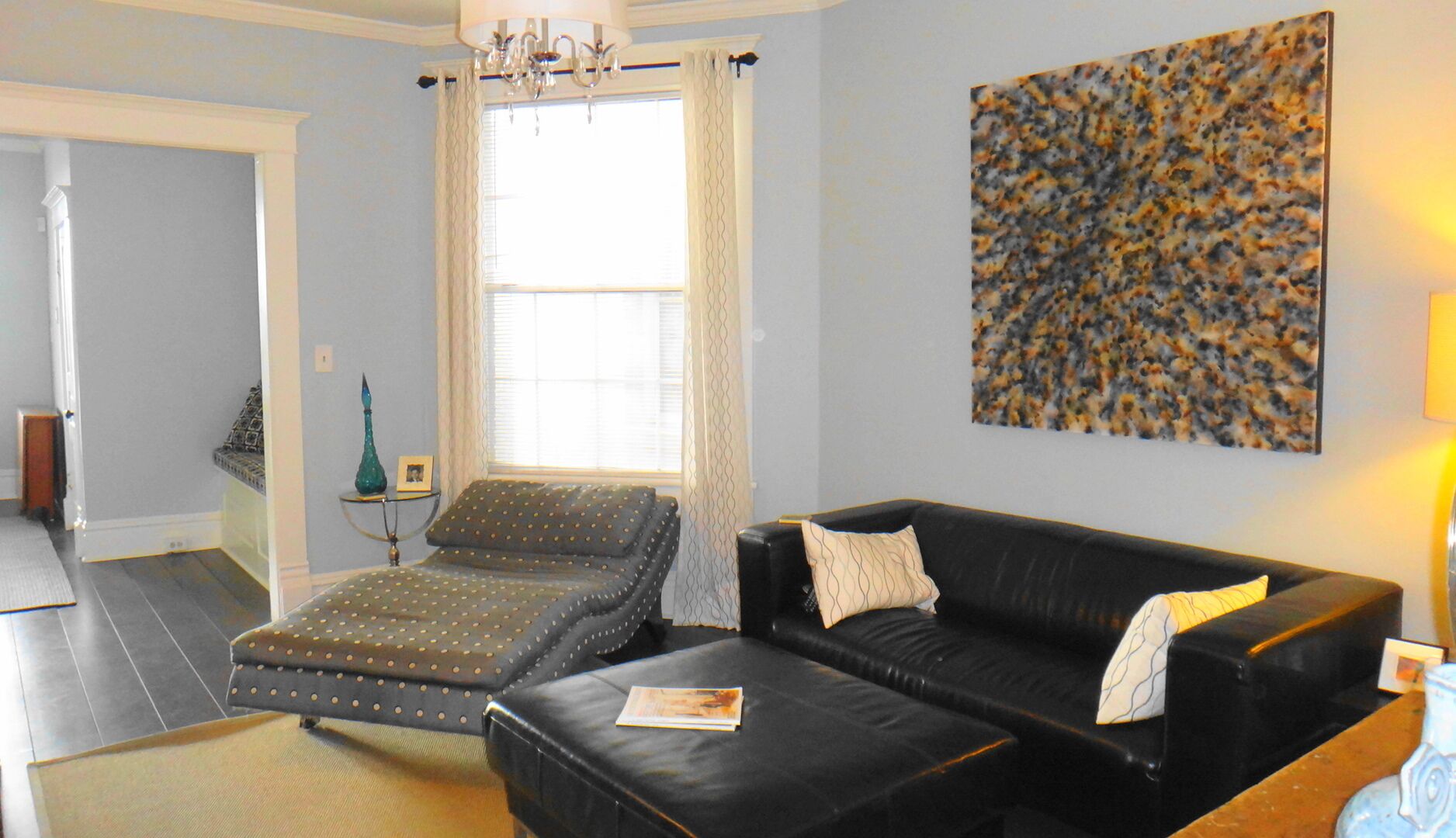 A living room with a black leather sofa, a patterned ottoman, a small side table with a vase, and a large abstract painting on the wall.