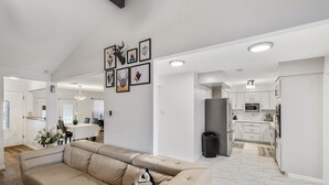 A spacious living room with a beige sofa, a dining area with a white table, and a kitchen with white cabinets and appliances. - Spacious 4BR Family Home, 6 Beds, Arcade, Game Room + Park Across Street (Watauga)