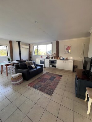 En moderne stue med sofa, spisebord med stole, et køkkenområde med mikroovn og et stort vindue, der giver naturligt lys.
