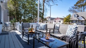 A deck with outdoor furniture, including chairs and a table, set against a backdrop of residential houses and trees. - Walk to the Beach! Firepit, Fully Fenced Yard, Grill, Bikes, N64, Pets/EV! (Ocean City)