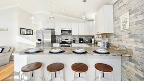 A modern kitchen with white cabinets, a marble countertop, and a wooden backsplash. There are three stools at the island and a coffee machine on the counter. - Walk to the Beach! Firepit, Fully Fenced Yard, Grill, Bikes, N64, Pets/EV! (Ocean City)