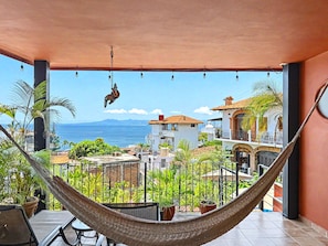 A hammock on a balcony overlooking a coastal view with buildings and vegetation. - Miguel's Mirador: Ocean-View Studio, Hammock Terrace, Heated Pool, Walkable (Puerto Vallarta)