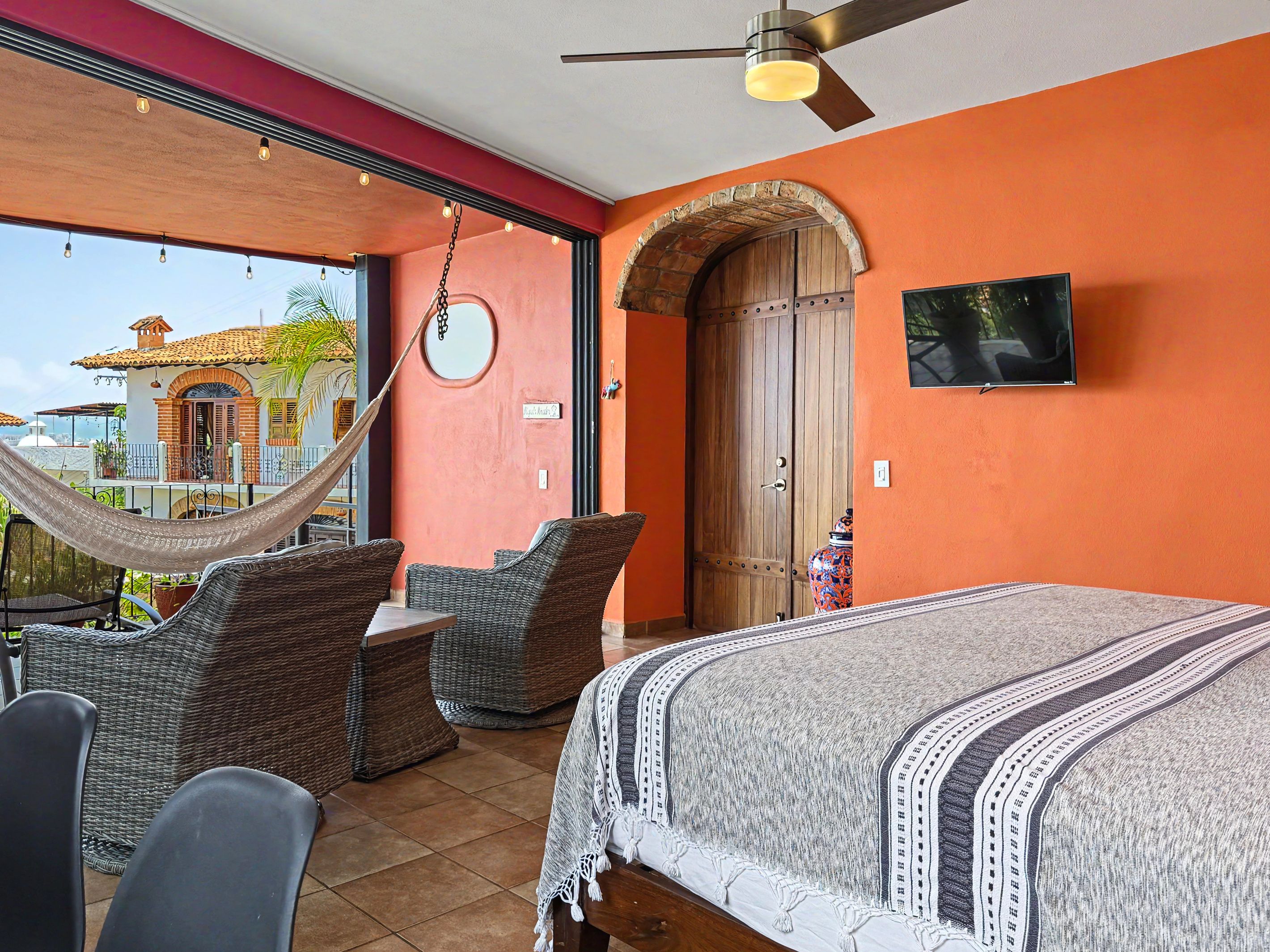 A bedroom with a bed, a hammock, wicker furniture, a television, and a large arched door.