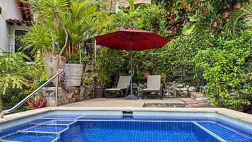 A swimming pool with blue tiles, surrounded by plants and a red umbrella.