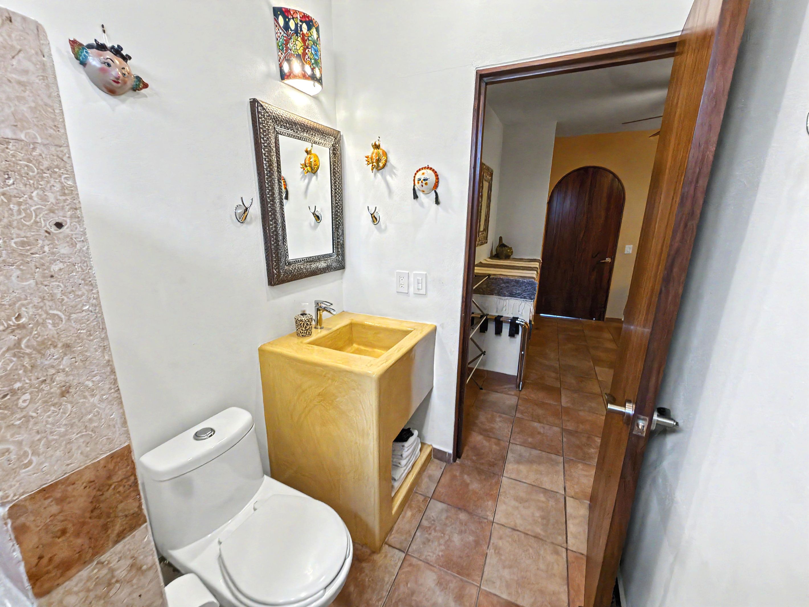 Un baño con inodoro, lavabo y espejo. Hay elementos decorativos en la pared y una puerta que da a otra habitación.