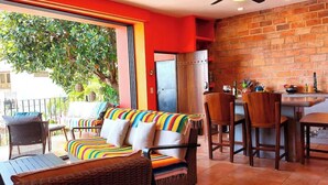 A cozy dining area with a wooden table and chairs, a colorful striped sofa, and a brick wall. - Ivan's Bella Vista: Top-Floor 2BR, Bay Views, Heated Pool, Walkable (Puerto Vallarta)