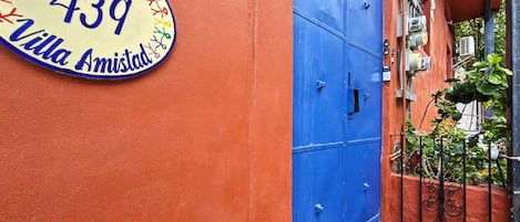 Een blauwe deur met een metalen hekwerk, een geel bord met 'Bienvenidos 439 Villa Amistad' op een rode muur, en een kleine trede die naar de deur leidt.
