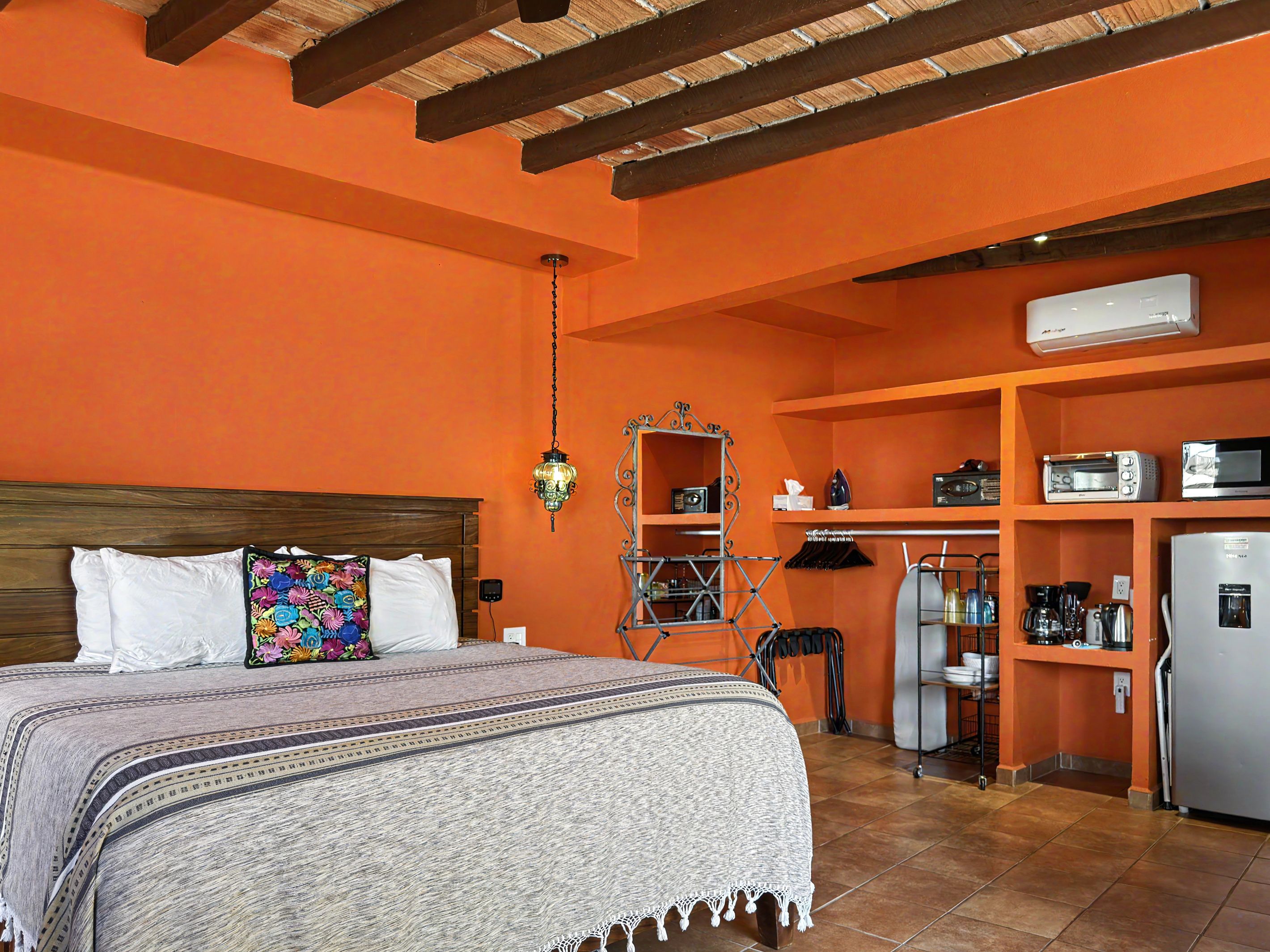 A bedroom with a bed, a colorful pillow, a hanging light fixture, and a kitchenette with a refrigerator, microwave, and coffee maker.