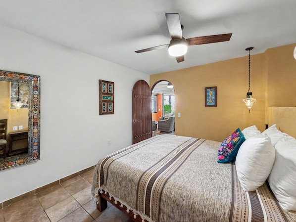 Un dormitorio con cama, ventilador de techo, espejo y puerta que da a otra habitación.