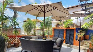 En udendørs terrasse med fletmøbler og store parasoller, omgivet af krukker med planter og udsigt til bygninger.