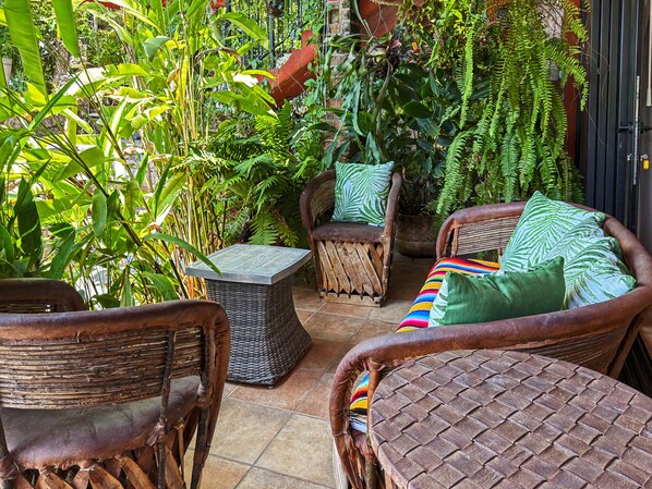 A patio area with wicker furniture, including a sofa and chairs, surrounded by lush green plants and ferns.