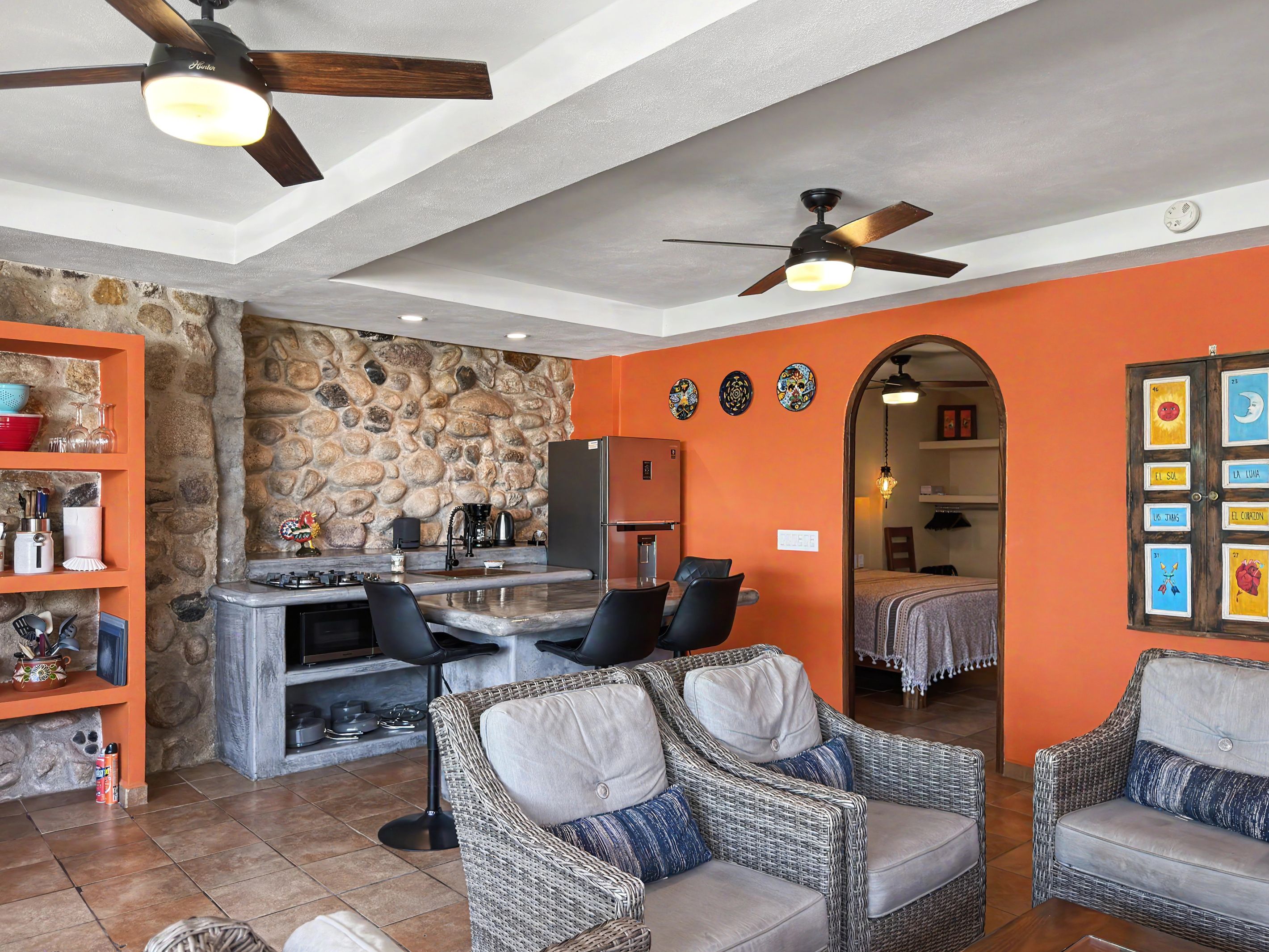 Una cocina moderna con pared de piedra, un comedor con muebles de mimbre y una sala de estar con paredes naranjas y ventiladores de techo.