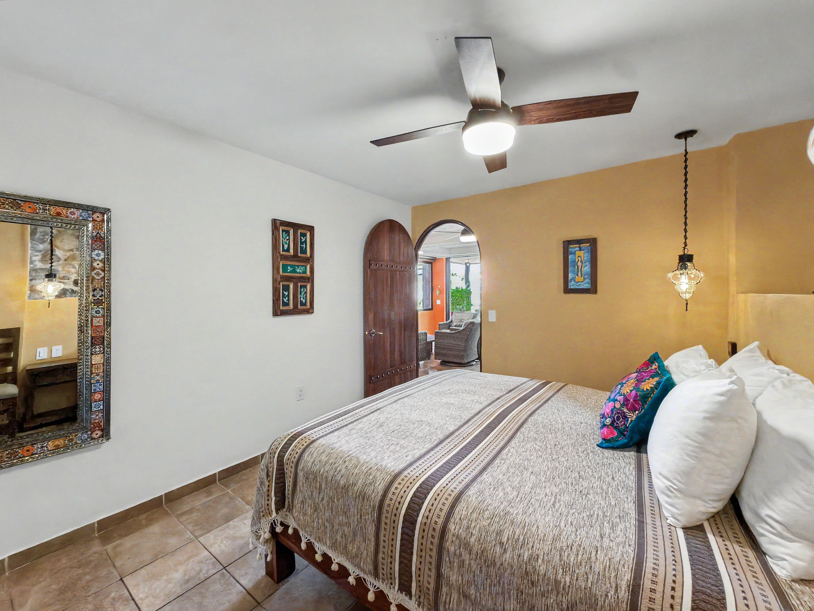 Un dormitorio con cama, ventilador de techo, espejo y puerta que da a otra habitación.