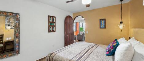 Un dormitorio con cama, ventilador de techo, espejo y puerta que da a otra habitación.