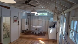Une cuisine compacte avec un réfrigérateur blanc, un évier et une cuisinière. On y trouve un coin repas avec une table et des chaises blanches. La pièce est dotée d’un plancher en bois et de murs blancs.
