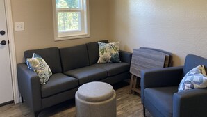 Sala de estar com sofá seccional cinza escuro, um banquinho redondo e uma poltrona azul. Há uma janela com vista para árvores e uma mesa pequena com tampo de madeira.