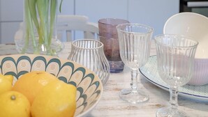 Une cuisine avec des meubles blancs, un pare-feu décoré, un vase rempli de tulipes roses, un bol de citrons et de la vaisselle transparente posée sur une table blanche.