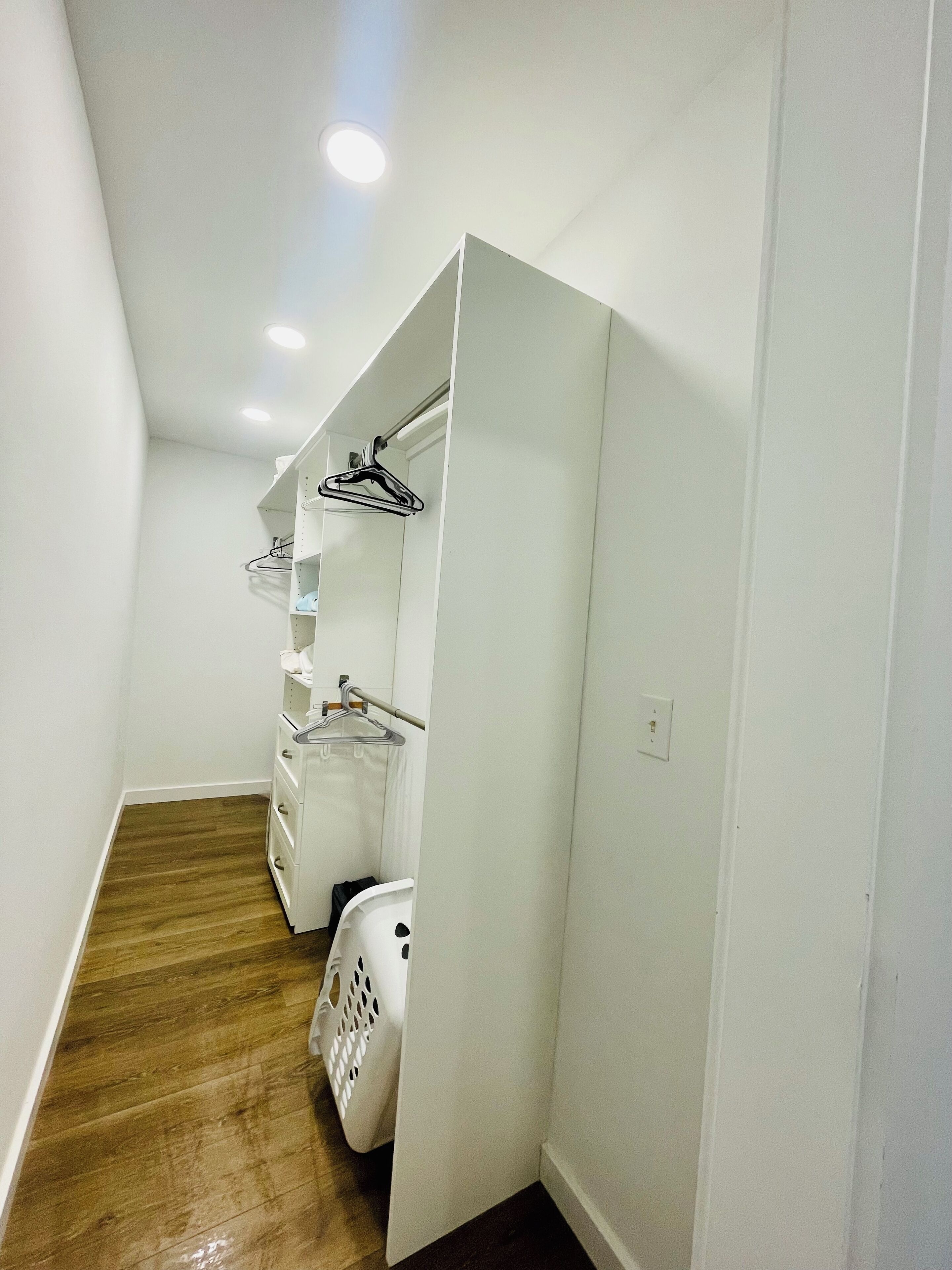 Un baño estrecho con paredes blancas, suelo de madera, una cesta de ropa blanca y un perchero con ropa.