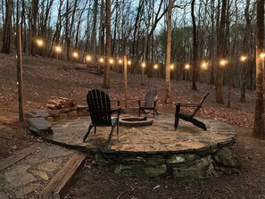 Un patio circolare in pietra con due poltrone Adirondack e un braciere, immerso in una foresta.
