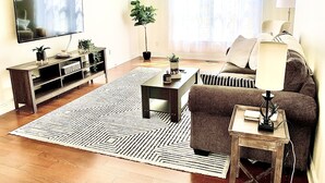A living room with a flat-screen TV, a sofa, a coffee table, a side table, a lamp, a potted plant, and a patterned rug. - The Relaxing Oaks (Columbus)