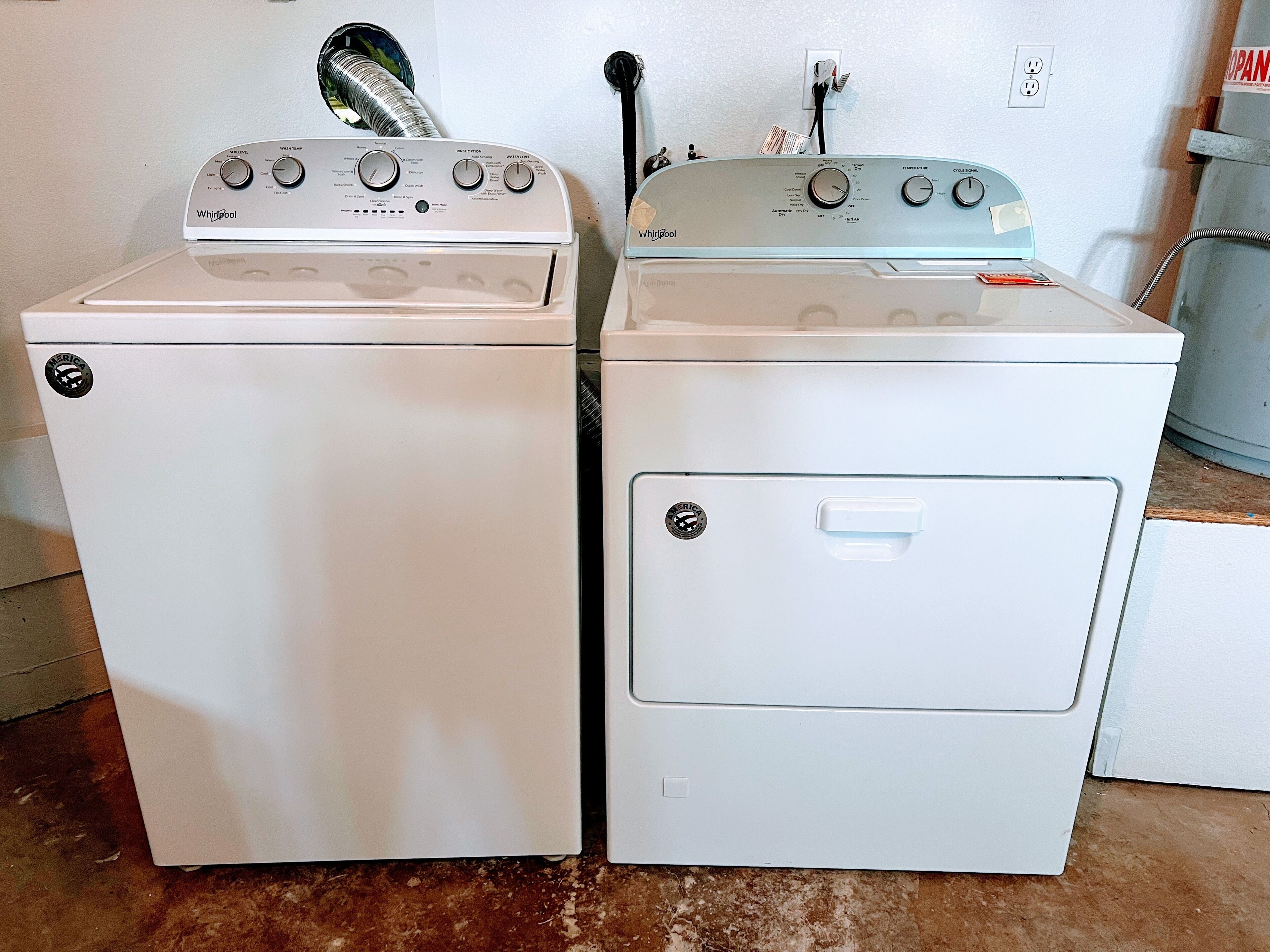 Two white Whirlpool washers with control panels, a power outlet, and a water heater on the wall.
