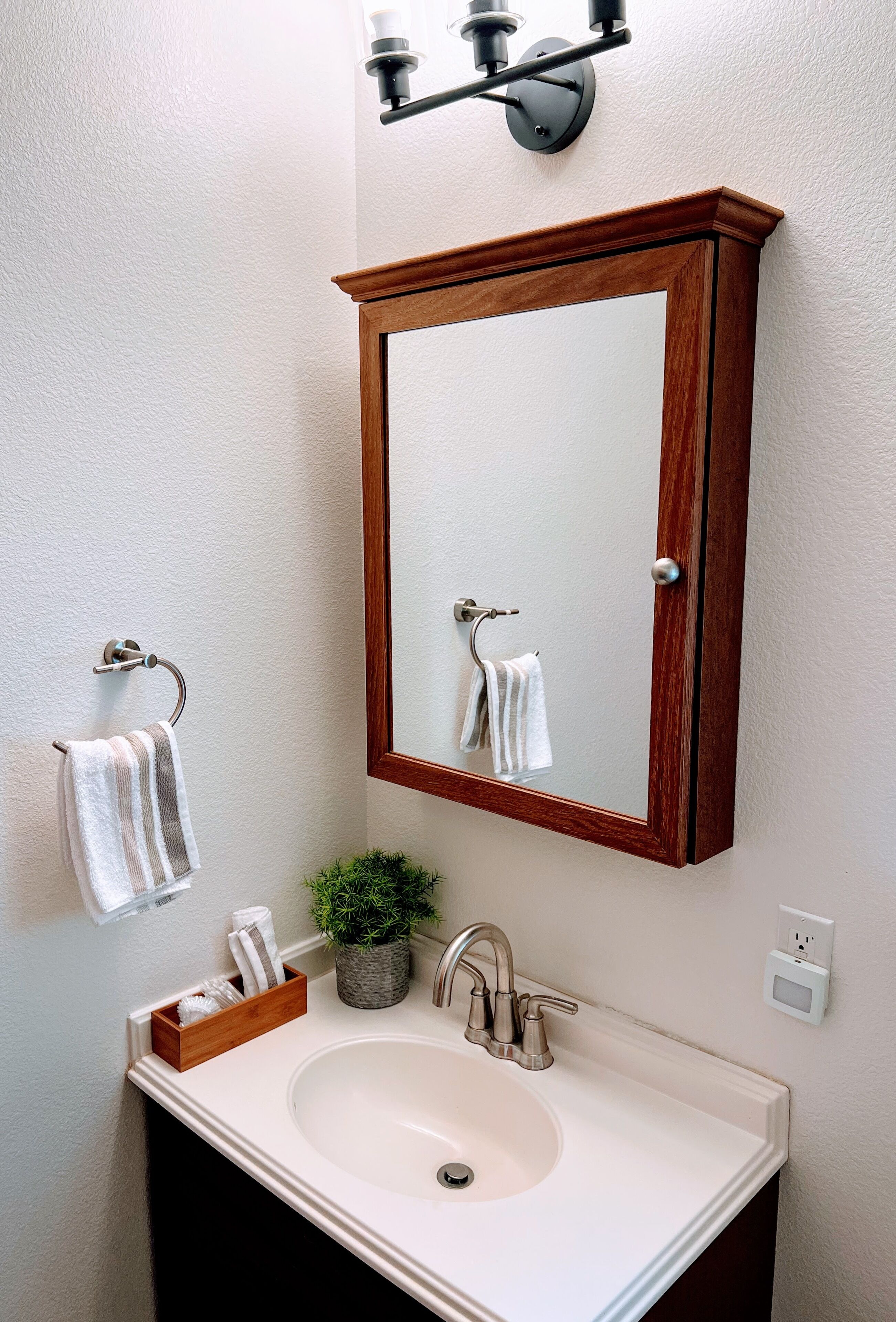 A bathroom with a wooden-framed mirror, a white sink, a towel rack, a potted plant, and a light fixture.