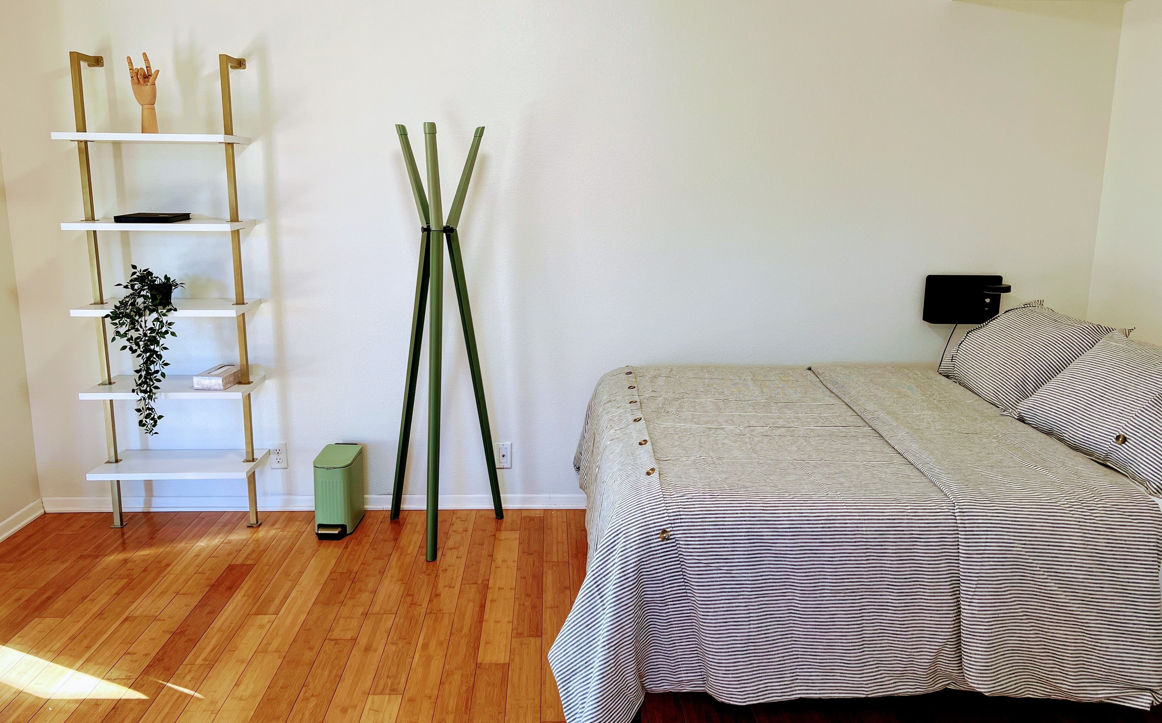 A bedroom with a bed, a bedside table, a lamp, a shelf with plants, and a trash bin.