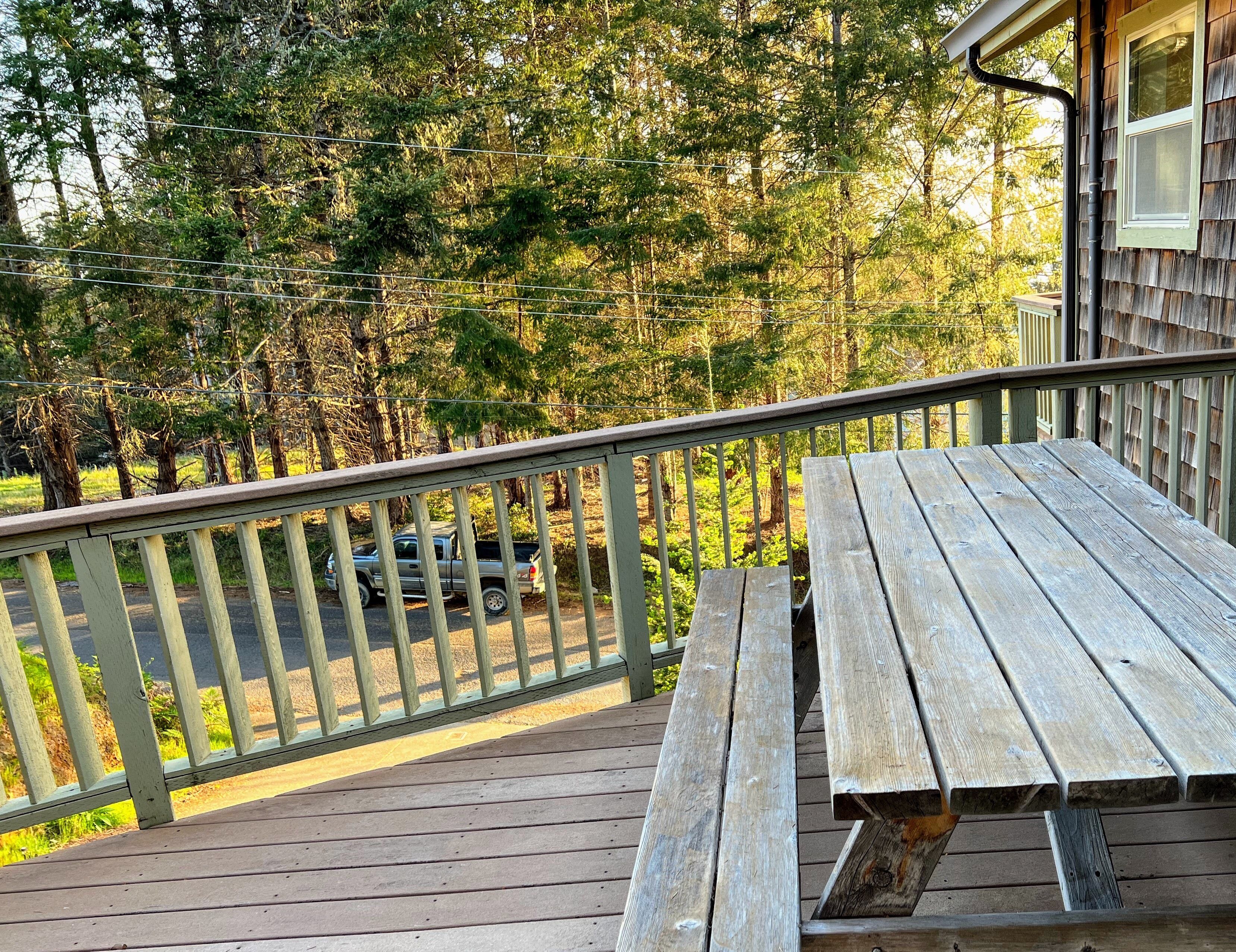 Une terrasse en bois avec une table de pique-nique et une rambarde, donnant sur une zone boisée où l’on aperçoit un véhicule garé au loin.