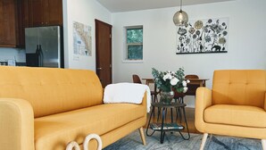 Een woonkamer met een bank, een klein krukje, een eettafel met stoelen, een koelkast en een muur met een bloemschilderij.