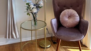 Een kamer met een raam, een vloerlamp, een stoel en een glazen tafel met een bloevas.