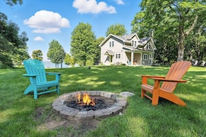 A house with a fire pit in the front yard, two Adirondack chairs, and a lake in the background.