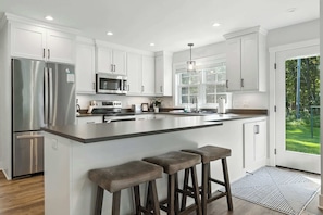 A modern kitchen with white cabinetry, stainless steel appliances, a large island with bar stools, and a view of a green outdoor area.