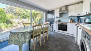 A modern kitchen with white cabinets, a dining table with chairs, and a large window. - A cozy cottage on the North Norfolk coast (Brancaster Saithe)