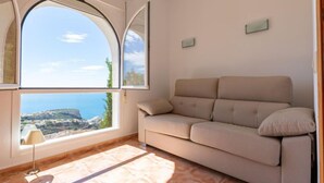 Ein Zimmer mit einem großen Bogenfenster, das einen schönen Ausblick bietet, einem beige farbenen Sofa, einem kleinen Tisch mit einer Lampe und einem Fliesenboden.