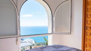 Ein Schlafzimmer mit einem großen Bett, Meerblick durch bogenförmige Fenster und einer Holztür.