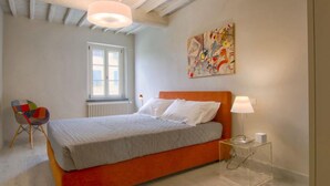 Ein Schlafzimmer mit einem Bett, einem Sessel, einem Fenster, einer Heizung, einem Bild und einem Nachttisch mit einer Lampe.