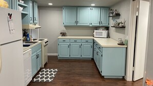 Une cuisine avec des meubles de couleur bleu clair, un réfrigérateur blanc, un four à micro-ondes et une cuisinière. Il y a un tapis sur le sol et une pancarte au mur sur laquelle on peut lire : « Quartiers du capitaine ».