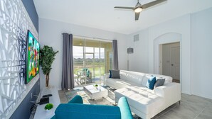 A modern living room with a white sofa, a coffee table, and a TV mounted on a white wall. There is a ceiling fan, a large window with curtains, and a view of the outdoors.