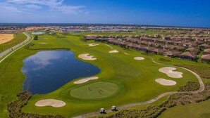 An aerial view of a golf course with sand bunkers, a pond, and residential housing in the background.