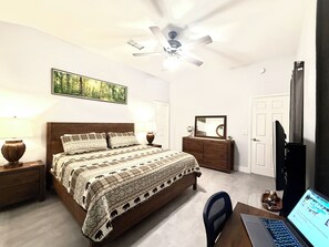 Une chambre à coucher avec un lit, un bureau avec un ordinateur portable, un ventilateur de plafond, une commode et une porte.