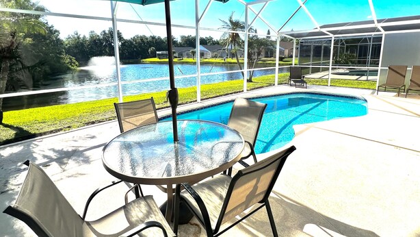 Área à beira da piscina com mesa e cadeiras de vidro, um grande guarda-sol e vista para um lago e vegetação.
