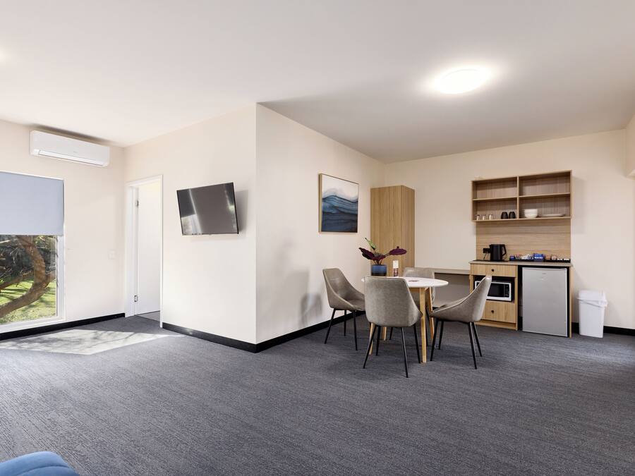 Una habitación de hotel moderna con un ventanal grande, un televisor de pantalla plana, una zona de comedor con mesa y sillas, una kitchenette y un refrigerador pequeño.