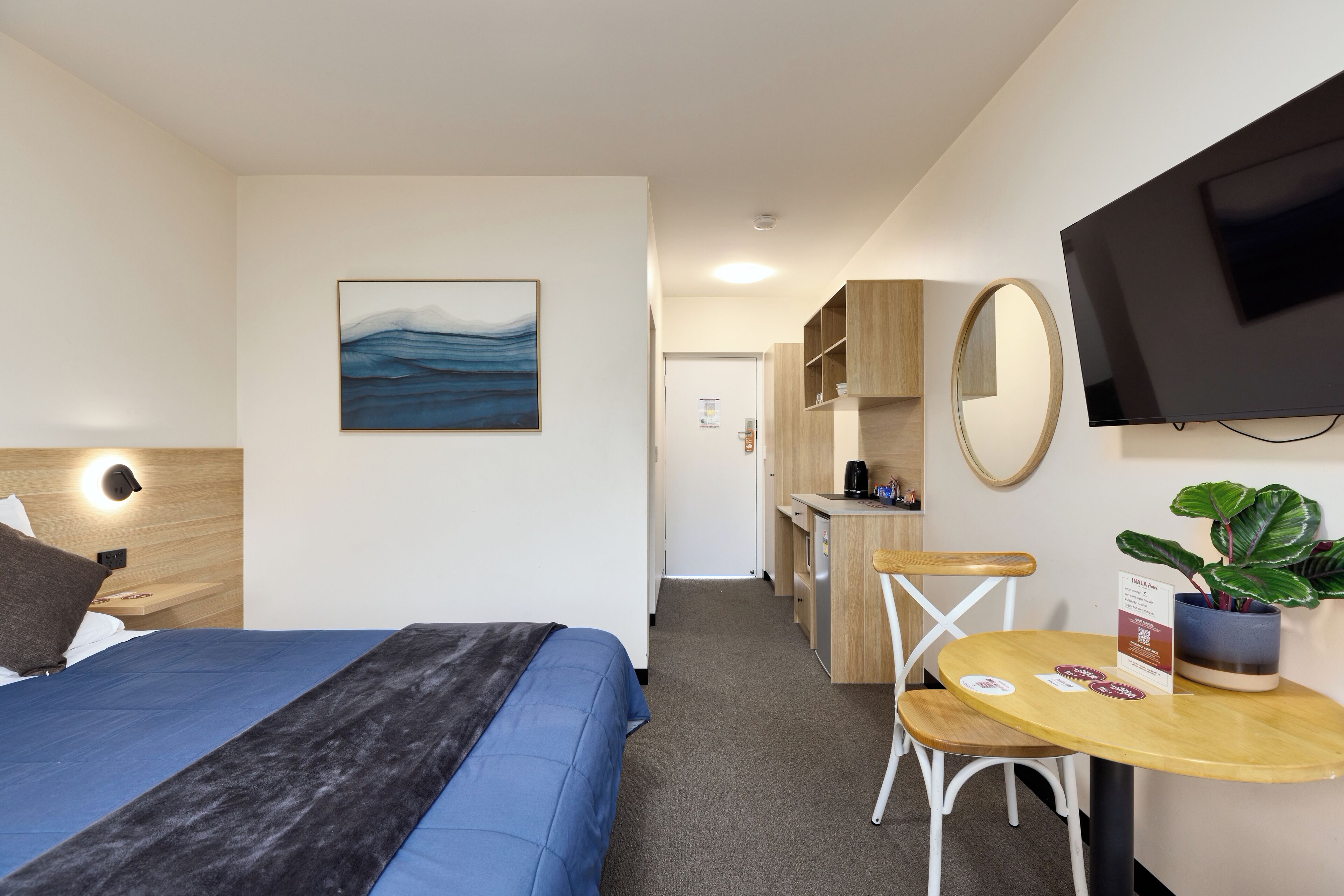 Quarto de hotel com cama, escrivaninha, cadeira, televisão, espelho e planta.