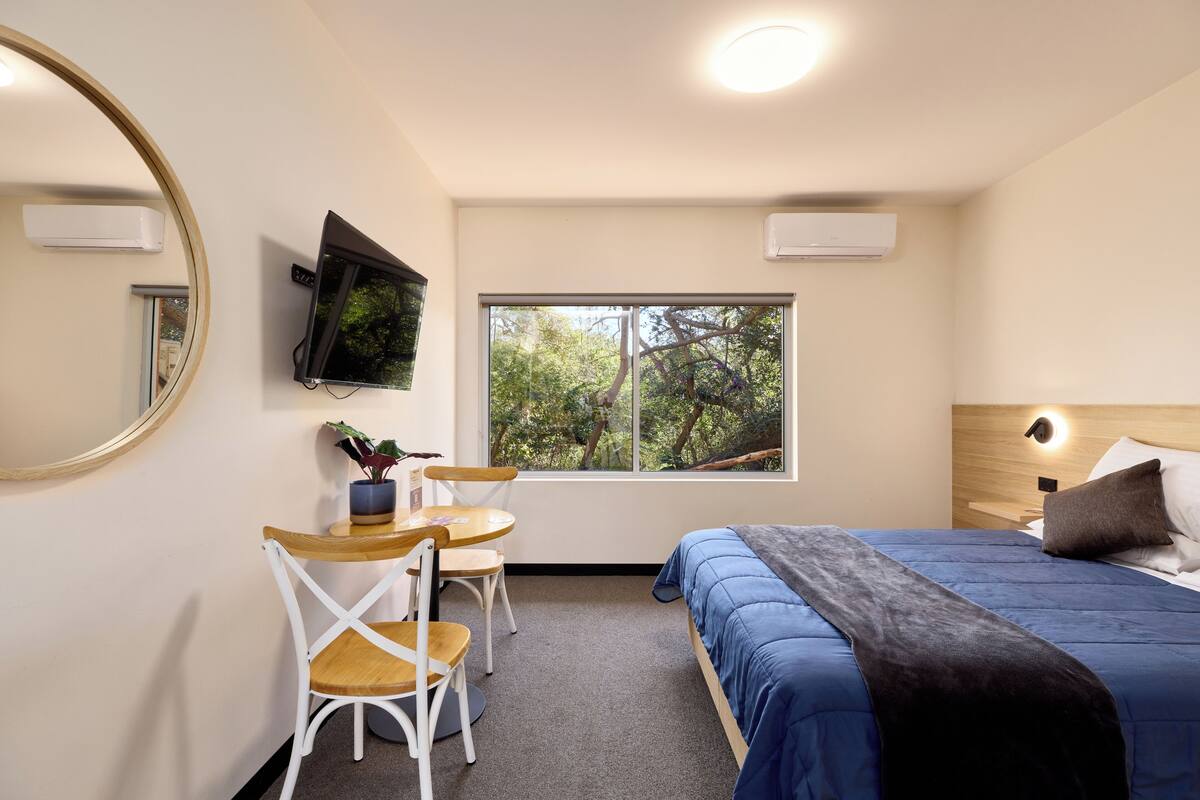 A bedroom with a bed, a desk, a chair, a window with a view of trees, and a mirror.