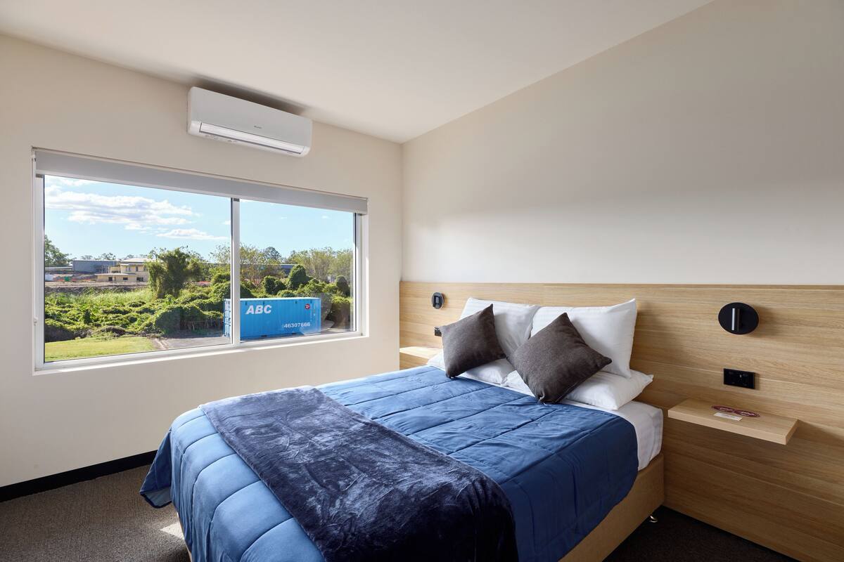 A hotel room with a large bed, a window with a view of greenery and buildings, a wooden headboard, and a small bedside table.