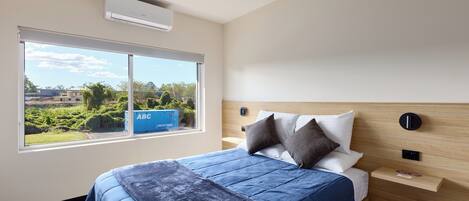 A hotel room with a large bed, a window with a view of greenery and buildings, a wooden headboard, and a small bedside table.