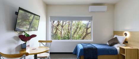 A small, modern bedroom with a bed, a TV mounted on the wall, a window with a view of trees, and a small table with chairs.
